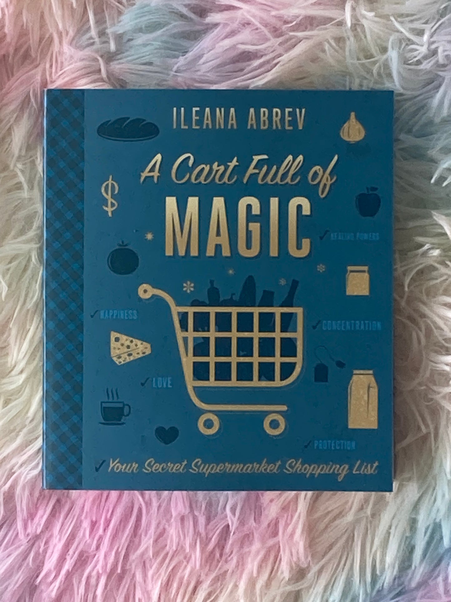 A book titled "A Cart Full of Magic: Your Secret Supermarket Shopping List" by Ileana Abrev is displayed on a fluffy, pastel-colored surface with shades of pink, purple, and blue. The book cover is teal blue with gold and dark blue illustrations of grocery items such as bread, garlic, cheese, milk, tea, and a coffee cup. A large shopping cart is centered on the cover, with words like “happiness,” “love,” “protection,” “concentration,” and “healing powers” surrounding it. The design suggests a magical theme