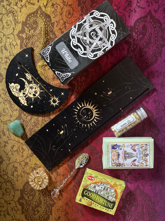 A witchcraft-themed kit is artfully arranged on a rich, gradient cloth that shifts from gold to red to magenta with a paisley pattern. Items include a box of white “Magic Mini Candles” with a pentacle design, a black moon-shaped Tarot card holder decorated with celestial artwork, a green aventurine crystal, a long tarot card holder with gold sun and moon illustrations, a small glass vial labeled “Jasmine” filled with Jasmine flowers, a bar of “Florida Water Soap,” a box of HEM “Good Fortune” incense cones, 
