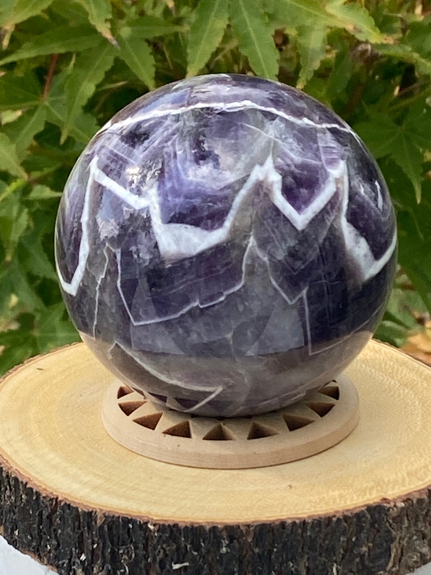 A polished amethyst sphere rests on a geometric wooden stand, placed atop a natural wood slice with bark edging. The sphere displays striking patterns of deep purple, lavender, and white in jagged, lightning-like bands. In the softly blurred background, green leaves add a natural, serene atmosphere to the scene.